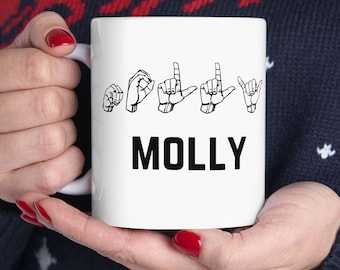 Pode incluir: Caneca de cerâmica branca com o nome "MOLLY" em letras pretas. Acima do nome, gestos com as mãos representando o nome em linguagem gestual. A caneca é segurada por uma pessoa que usa um suéter azul escuro com um padrão de renas vermelhas e brancas.