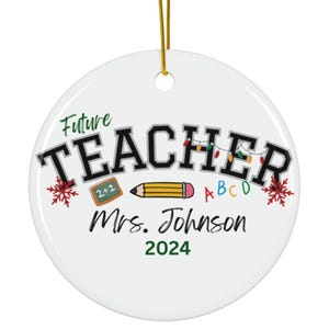 以下が含まれることがあります： 金色の紐で吊るされた丸い白いセラミック製のオーナメント。Future TEACHER、Mrs. Johnson、2024という文字が書かれています。2+2と書かれた小さな黒板、黄色い鉛筆、カラフルなクリスマスライト、赤い雪の結晶のグラフィックが描かれています。