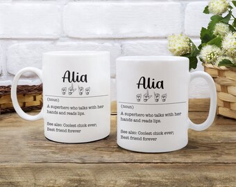 Pode incluir: Duas canecas de cerâmica branca com o nome "Alia" impresso nelas. As canecas têm um contorno preto e o nome está escrito em letra cursiva. As canecas também têm uma definição do nome em linguagem de sinais. A definição diz: "Uma super-heroína que fala com as mãos e lê os lábios. Veja também: A garota mais legal de todos os tempos. Melhor amiga para sempre."