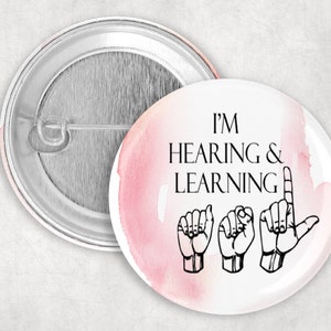 May include: A white button with a pink watercolor background. The button says "I'm Hearing & Learning" in black text with a sign language depiction of the phrase.