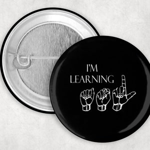 Peut inclure: Bouton noir avec du texte blanc qui dit "J'APPRENDS" et trois signes de main blancs représentant les lettres "L", "E" et "A" en langue des signes française.