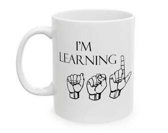 Pode incluir: Uma caneca de cerâmica branca com o texto preto I'M LEARNING acima de um desenho de linha preta de três mãos realizando os sinais manuais da língua de sinais americana para as letras A, S e L, o acrónimo para esta língua de sinais.