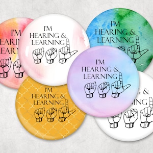 Peut inclure: Six boutons ronds avec un fond aquarelle dans différentes couleurs. Chaque bouton présente le texte "Je suis sourd et j'apprends" avec une représentation en langue des signes de la phrase.