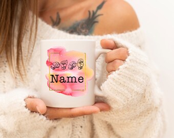 Pode incluir: Caneca de cerâmica branca com um design em aquarela em tons de rosa e amarelo, emoldurada por um quadrado dourado. A caneca exibe formas de mãos em ASL e a palavra "Name" em preto.
