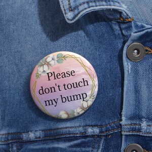 Puede incluir: Un botón redondo con un fondo de acuarela en tonos rosa y morado. El botón presenta un marco geométrico dorado con flores de algodón blancas y hojas verdes. El texto "Please don't touch my bump" está escrito en negro.