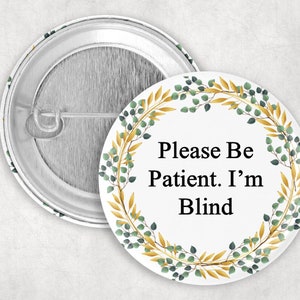 May include: White button with a gold and green wreath design. The text "Please Be Patient. I'm Blind" is written in black inside the wreath.