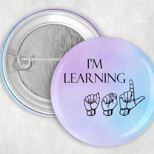 Peut inclure: Un bouton rond avec un fond dégradé bleu clair et violet. Le bouton présente le texte "I'M LEARNING" en noir avec trois signes de main noirs représentant les lettres "L", "E" et "A" en langue des signes américaine.