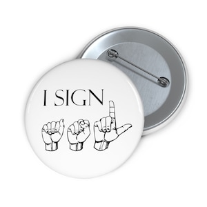 May include: White button with the text "I SIGN" in black lettering and a graphic of the sign language alphabet for the letters "I", "S", "I", "G", "N".