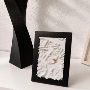 Può includere: Una cornice nera rettangolare contenente una mappa in rilievo tridimensionale bianca di una planimetria urbana. La mappa presenta blocchi di edifici bianchi in rilievo e una piccola icona a forma di cuore rosso su una delle strutture.