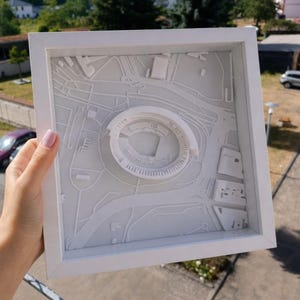 May include: A white framed 3D wall art piece featuring a monochromatic relief map of a city area. The center displays a circular stadium structure surrounded by raised topographical lines representing streets, buildings, and infrastructure in white.