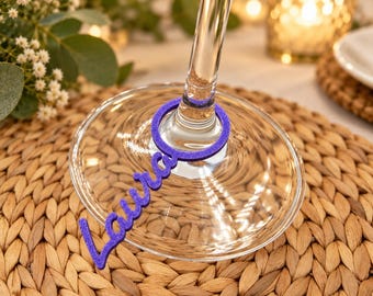 May include: A purple glittery acrylic wine glass charm featuring the name Laura in a cursive script font. The circular charm fits around the stem of a clear glass, resting on the base of the glass with a textured, sparkling finish.