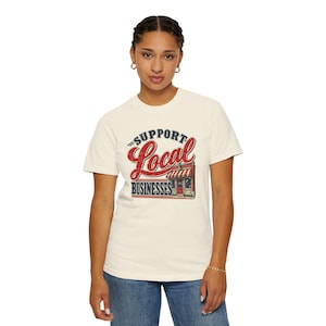 May include: A cream-colored short-sleeve t-shirt featuring a graphic print with the text SUPPORT LOCAL BUSINESSES in red and dark blue. The design includes a small storefront illustration with a striped awning and a distressed, vintage-style finish.
