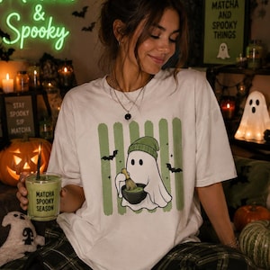 Peut inclure: Un t-shirt blanc à manches courtes avec un motif représentant un fantôme portant un bonnet vert, fouettant du matcha vert dans un bol noir. Le design inclut des rayures verticales vertes et des chauves-souris noires.