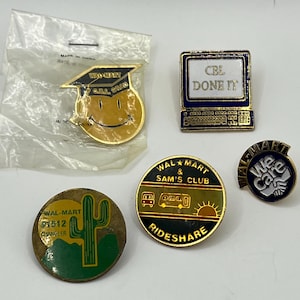May include: Five metal lapel pins featuring various designs: a yellow smiley face wearing a graduation cap, a computer screen with the text CBL DONE IT, a round pin with a cactus and text WAL-MART 1512 CHANDLER, a RIDESHARE pin, and a WE CARE pin.
