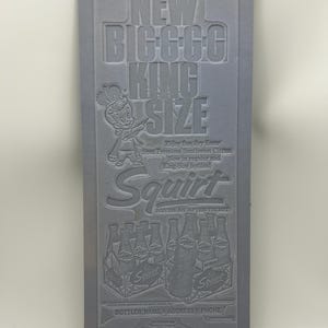 May include: A rectangular gray metal printing plate featuring embossed text and graphics for Squirt soda. The design includes the text NEW BIGGGG KING SIZE, a cartoon character wearing a crown, and an illustration of multiple soda bottles.