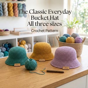 Könnte beinhalten: Drei gehäkelte Fischerhüte in Petrol, Senfgelb und Lavendel, jeweils mit einem Holzschildchen mit der Aufschrift Small, Medium oder Large. Der Text lautet The Classic Everyday Bucket Hat All three sizes Crochet Pattern. Häkelnadel und Garn liegen bei.