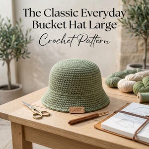 May include: A sage green crochet bucket hat with a small rectangular wooden tag labeled LARGE. The text The Classic Everyday Bucket Hat Large Crochet Pattern is visible above the hat, alongside metal scissors, a wooden crochet hook, and yarn.