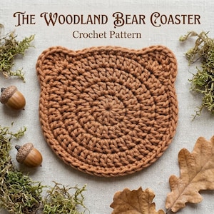May include: A brown crochet coaster shaped like a bear head with rounded ears. The text above reads The Woodland Bear Coaster Crochet Pattern. The coaster is surrounded by green moss, two brown acorns, and a dried oak leaf on a light fabric.