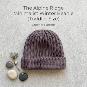 May include: A purple crochet beanie with a ribbed texture and a folded brim. The text above reads: The Alpine Ridge Minimalist Winter Beanie (Toddler Size) Crochet Pattern. The beanie is displayed with a small pine sprig and three smooth stones.