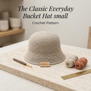 Puede incluir: Un sombrero de pescador de ganchillo beige con una pequeña etiqueta de madera que dice SMALL, acompañado de un ganchillo de madera, un ovillo de lana beige, un ovillo de lana terracota y una regla de metal. Texto: The Classic Everyday Bucket Hat small Crochet Pattern.