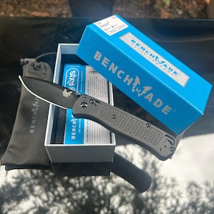 Könnte beinhalten: Ein schwarzes Klappmesser mit strukturiertem grauem Griff und Benchmade-Logo auf der Klinge. Es liegt neben der blau-weißen Produktverpackung mit der Aufschrift Benchmade Bugout und einem schwarzen Stoffbeutel mit dem Markennamen Benchmade.