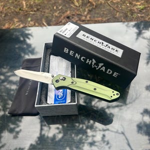 Peut inclure: Un couteau pliant Benchmade 940-1 Osborne avec un manche vert clair et une lame argentée, posé sur sa boîte grise ouverte. L'emballage noir présente le logo et le texte Benchmade, accompagné d'une pochette de rangement en tissu noir et d'inserts.