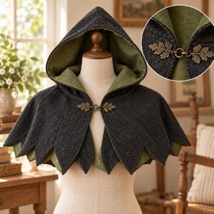 May include: A hooded capelet featuring a dark charcoal gray exterior and a sage green interior lining. The hem is cut into a series of pointed leaf-like shapes. A bronze-toned metal clasp shaped like two oak leaves secures the front closure.