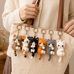 May include: Six crocheted cat keychains hanging in a row. Each cat features a different color pattern including brown tabby, calico, black and white, orange tabby, gray, and solid white. Each keychain has a metal ring and a small dangling cat figure.