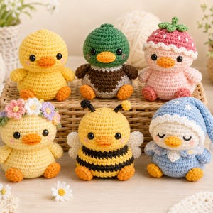 May include: Six small crocheted duck figures arranged in two rows. The top row features a yellow duck, a mallard-style duck, and a pink duck wearing a strawberry hat. The bottom row includes a duck with a flower crown, a bee-themed duck, and a sleeping duck.