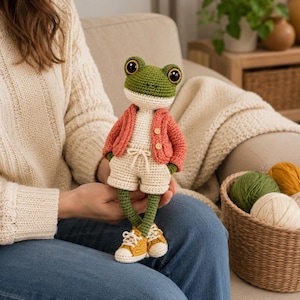Puede incluir: Una muñeca rana de ganchillo con grandes ojos negros, que lleva una rebeca color coral con dos botones pequeños, pantalones cortos de color crema con cordón y zapatillas amarillas y blancas. La rana tiene cabeza y extremidades verdes y pecho crema.