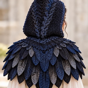 May include: A hooded crochet capelet featuring a layered feather-like design in shades of dark navy, royal blue, and charcoal grey. The hood and shoulder area are constructed with intricate stitch patterns, creating a textured, plumage-inspired look.