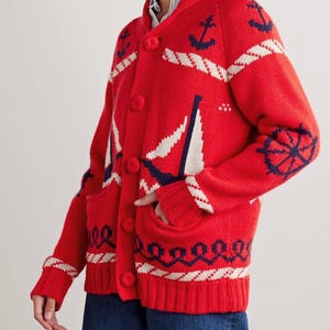 May include: A bright red knit cardigan featuring a nautical theme with navy blue anchors, ship wheels, and white sailboat motifs. The sweater has a shawl collar, long sleeves with ribbed cuffs, a button-front closure, and two front patch pockets.