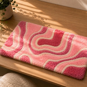Könnte beinhalten: Ein rechteckiger getufteter Teppich mit einem wellenförmigen, abstrakten Muster in den Farben Pink, Hellrosa und Creme. Der Teppich hat eine dicke, strukturierte Floroberfläche mit geschwungenen, fließenden Linien über die gesamte Länge.