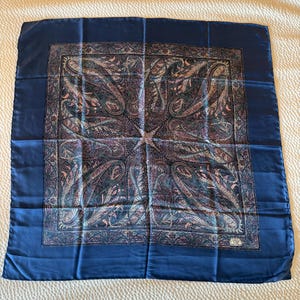 May include: A square silk scarf featuring a solid navy blue border and a central panel with a detailed paisley pattern in shades of blue, purple, and pink. A small white rectangular label in the bottom right corner reads Liberty of London Made in England.