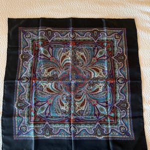 May include: A square scarf featuring a black border and a central intricate paisley pattern in shades of blue, red, green, and white. The design includes symmetrical floral motifs and geometric shapes. The word LIBERTY is printed in the bottom corner.