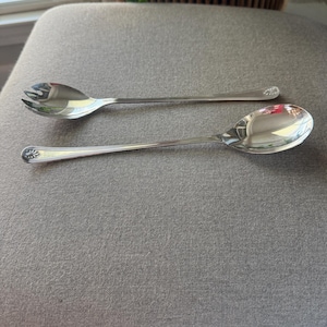 May include: A pair of silver-toned metal serving utensils consisting of a salad fork with three tines and a matching solid spoon. Both pieces feature a polished finish and a decorative shell-like motif embossed at the end of the slender handles.