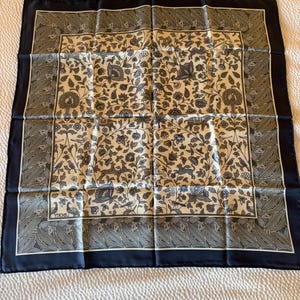 May include: A square scarf featuring a cream-colored center with a dark blue and grey botanical print of flowers, birds, and animals. The scarf has a wide dark blue border with a leaf pattern and the word LIBERTY printed in the bottom left corner.