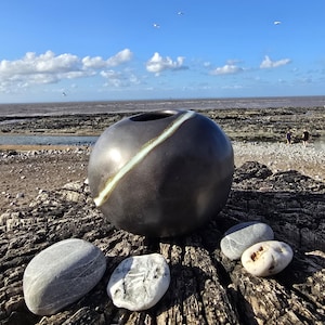May include: A spherical dark charcoal ceramic vase with a smooth matte finish and a single diagonal light green streak running across its surface, positioned alongside three smooth natural river stones on a weathered wooden surface.