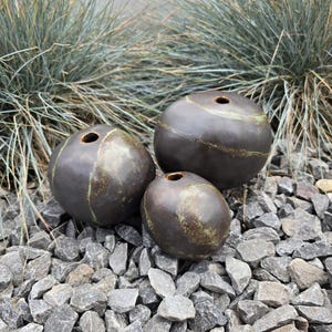 May include: Three spherical ceramic vases with a dark charcoal finish and subtle, irregular green-toned streaks. Each vase features a small, circular opening at the top, showcasing a smooth, rounded form with a slightly textured, organic appearance.