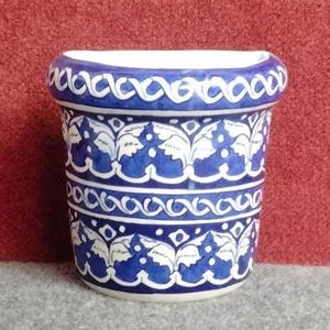 Puede incluir: Una maceta de pared de cerámica con forma semicircular y parte posterior plana. El exterior está decorado con un patrón pintado a mano de hojas blancas estilizadas y motivos geométricos de ondas sobre un fondo azul intenso y base blanca.