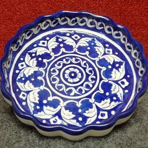 May include: A round ceramic baking dish with a scalloped edge, featuring a deep blue and white hand-painted floral and geometric pattern. The interior displays a central flower motif surrounded by intricate leafy scrolls and a border design.