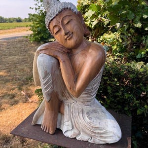 Puede incluir: Una estatua de Buda de madera tallada a mano con un acabado de grano de madera marrón natural en la parte superior del cuerpo y un acabado blanco desgastado en la túnica inferior y el cabello. La figura está en una pose sentada.