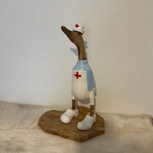 May include: A carved wooden duck figurine standing on a flat wooden base. The duck is painted to resemble a healthcare worker, wearing a white nurse cap with a red cross, a light blue tunic with a red cross emblem, and white clogs on its feet.