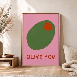 Könnte beinhalten: Ein rechteckiger Wandkunstdruck mit einem einfarbigen rosa Hintergrund und einer großen, mittig platzierten grünen Olivenform. In der Olive befindet sich ein kleines, rotes herzförmiges Piment. Unter der Olive steht der Text OLIVE YOU in roten Buchstaben.