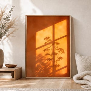 Peut inclure: Une impression d'art mural rectangulaire dans un cadre fin présentant un fond orange profond avec la silhouette sombre d'une plante projetée par l'ombre d'une fenêtre. La composition affiche un motif d'ombre en grille sur la forme botanique.