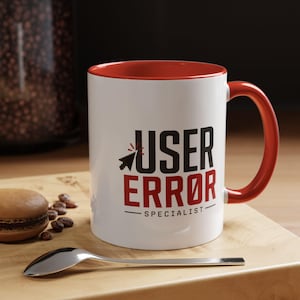 May include: A white ceramic mug with a red interior and handle. The front features a black pixelated cursor icon next to the text USER in black, followed by ERROR in red, and the word SPECIALIST in black underneath with a horizontal line.