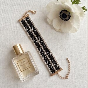 May include: A beaded bracelet featuring a geometric pattern of small black and white beads arranged in two parallel columns, finished with gold-toned metal end bars and a lobster clasp closure with an adjustable chain extension.