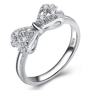 May include: A silver-toned ring featuring a bow-shaped centerpiece adorned with multiple small, clear, round-cut stones. The band is polished and includes an S925 hallmark engraving on the inner surface, indicating sterling silver composition.
