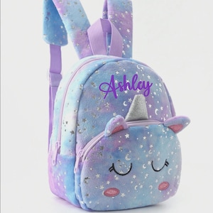 May include: A plush backpack featuring a blue and purple tie-dye pattern with silver stars and moons. The front pocket displays a unicorn face with a silver horn, closed eyes, and pink cheeks. The name Ashley is printed in purple script on the top.