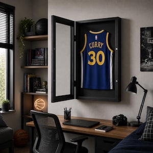 Peut inclure: Une vitrine en bois noir avec une porte vitrée fixée au mur, contenant un maillot de basket bleu à bordures jaunes, avec le nom CURRY et le numéro 30 imprimés en jaune au dos. La vitrine est ouverte, révélant un intérieur noir.